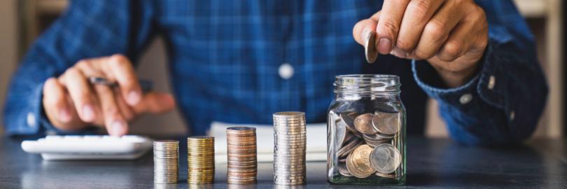 Ein Mann füllt ein Glas mit Münzen. Neben ihm sind Münzen, die kleine Säulen bilden.