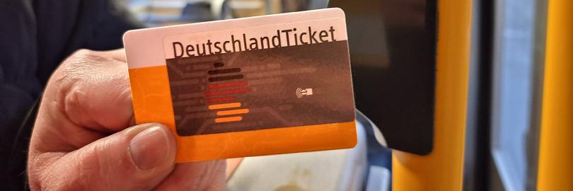 Eine Hand hält ein Deutschlandticket neben einen Stopp-Knopf an der Tür einer Straßenbahn