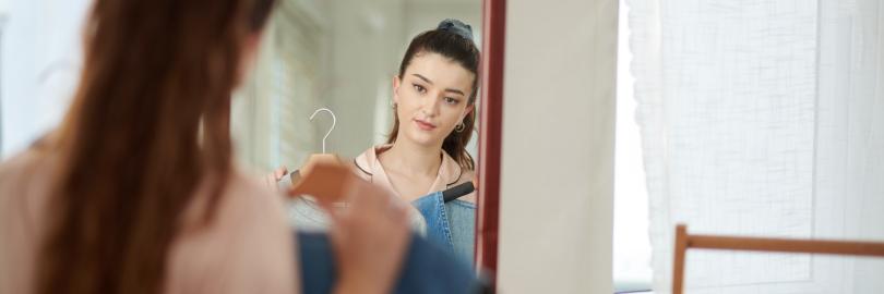 Eine junge Frau mit braunem Haar steht vor einem Spiegel und hält verschiedene Kleidungsstücke auf Bügeln vor ihren Körper. Ihr Gesicht ist in der Spiegelreflexion deutlich zu sehen, während sie prüfend in den Spiegel blickt.