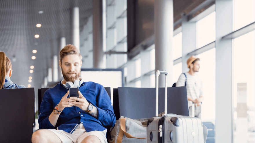 junger Mann mit Gepäck sitzt in einer Abflughalle und schaut auf sein Smartphone