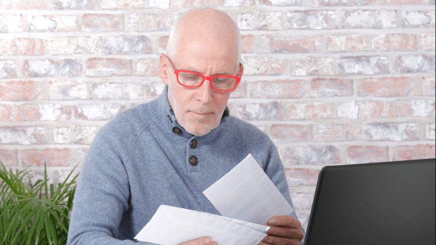 älterer Mann mit Glatze und roter Brille sieht seine Post mit ernster Miene durch