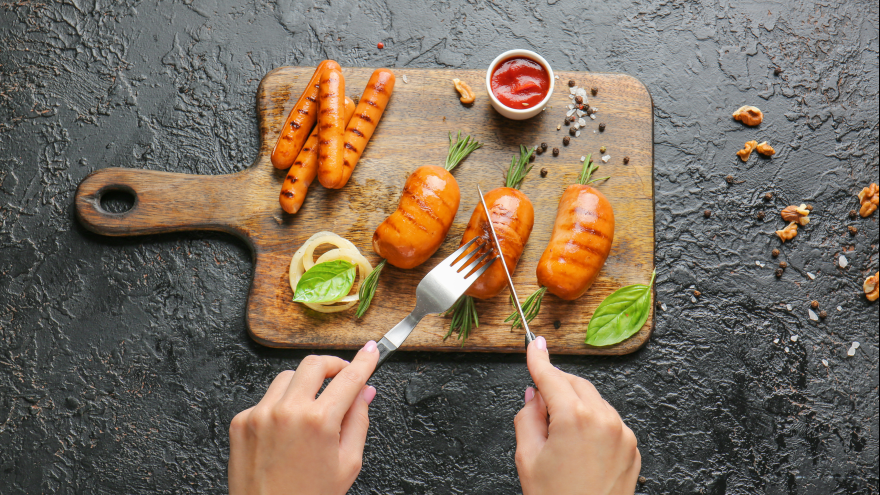 ein Holzbrett mit vegetarischen Würstchen, Ketchup und etwas Gemüse, zwei Hände halten Besteck
