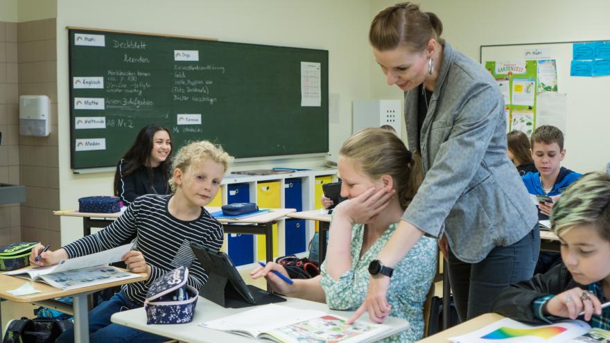 Klassenraum mit Lehrerin, die einer Schülerin etwas an ihrem Platz zeigt.