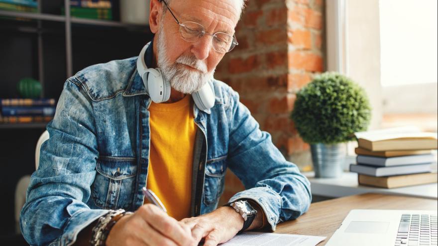 älterer Mann mit weißem Haar, Brille, Bart und Kopfhörern arbeitet am Schreibtisch