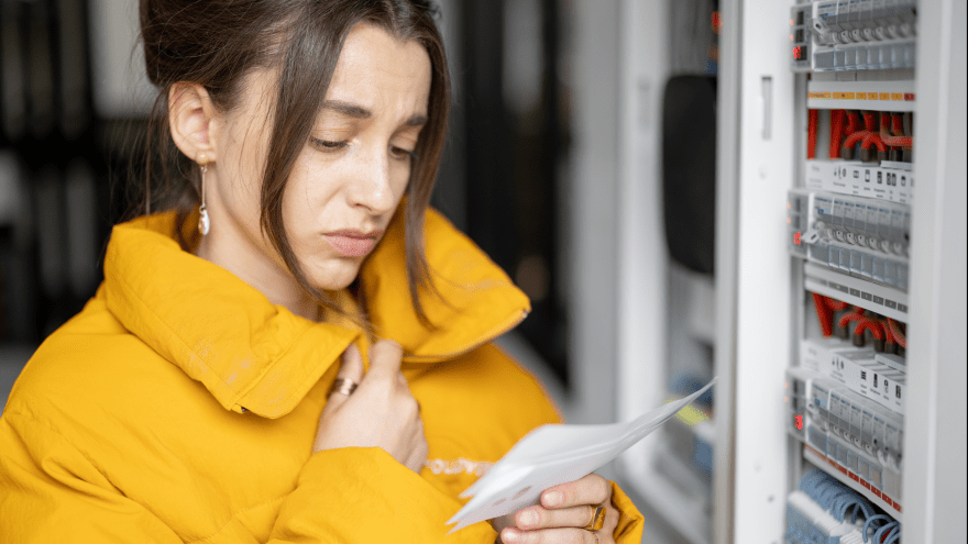 Eine Frau steht vor einem Stromkasten und liest eine Abrechnung durch. 