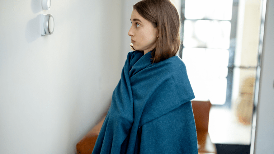 junge Frau, in eine blaue Decke gehüllt, blickt ernst auf einen Heizungsregler.