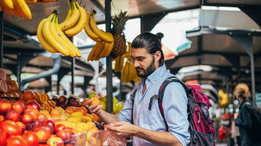 junger Mann mit Bart und Rucksack am Obststand in Markthalle