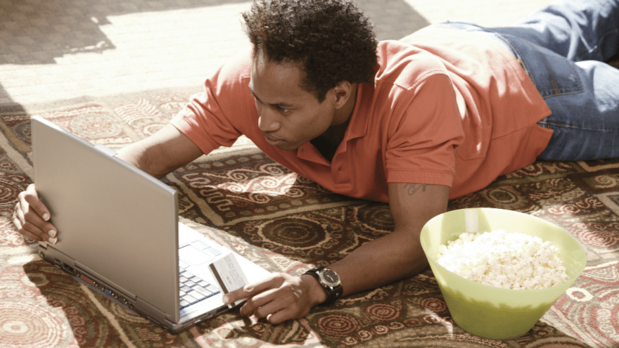 junger, dunkelhäutiger Mann liegt mit Laptop und Kreditkarte auf einem Teppich, schaut auf den Bildschirm