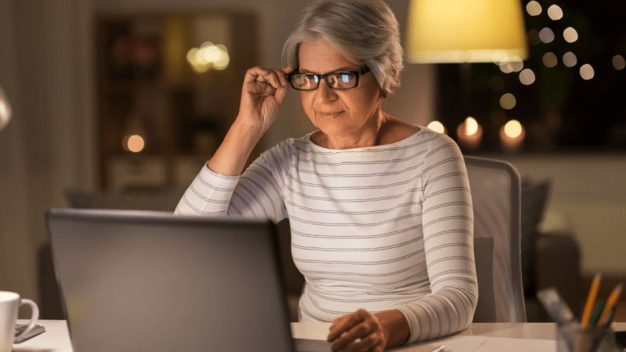 Frau im Rentenalter mit Brille sitzt am Laptop, abendliche Wohnzimmeratmosphäre