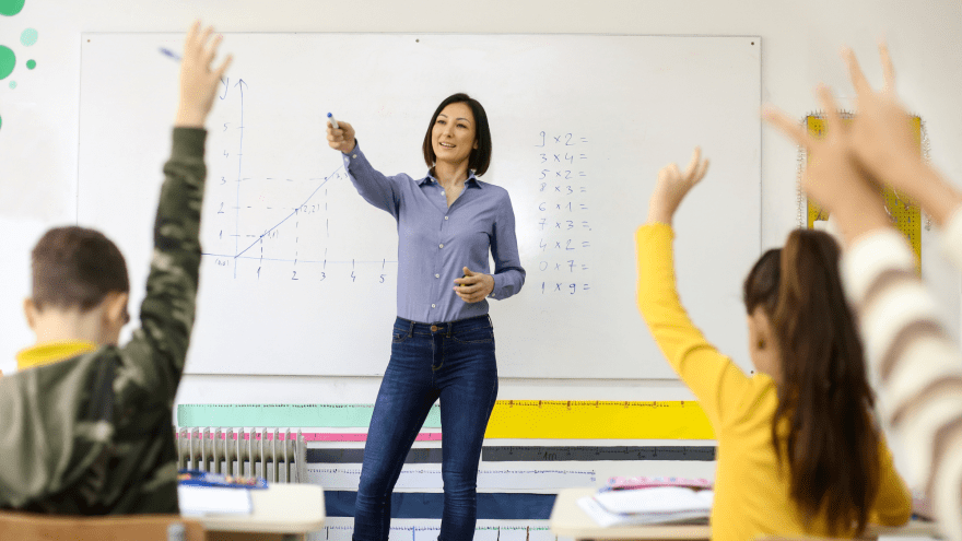 Lehrerin steht vor einer Klasse. Die Schulkinder zeigen auf.