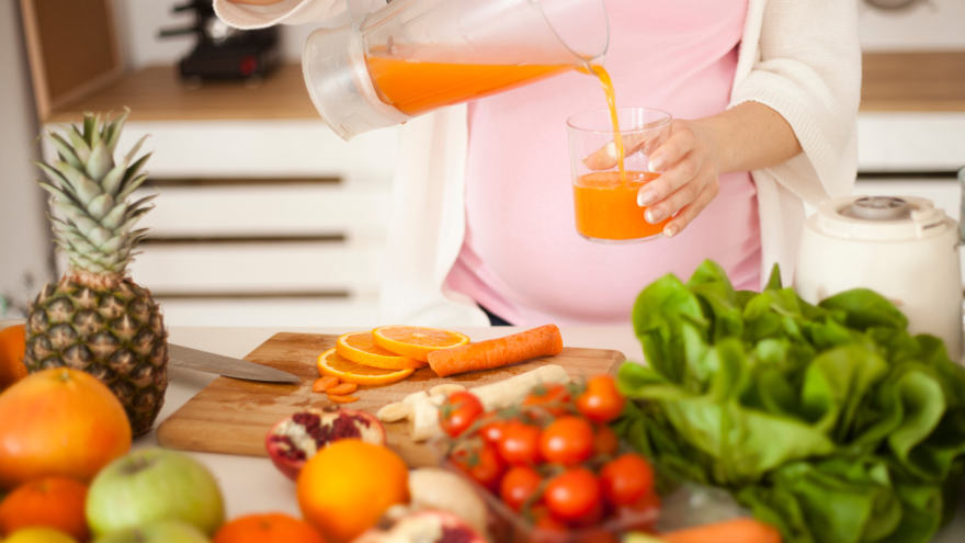 Eine schwangere Frau steht vor einer Kucheninsel voller Obst und Gemüse, sie schenkt sich ein Glas Karottensaft ein