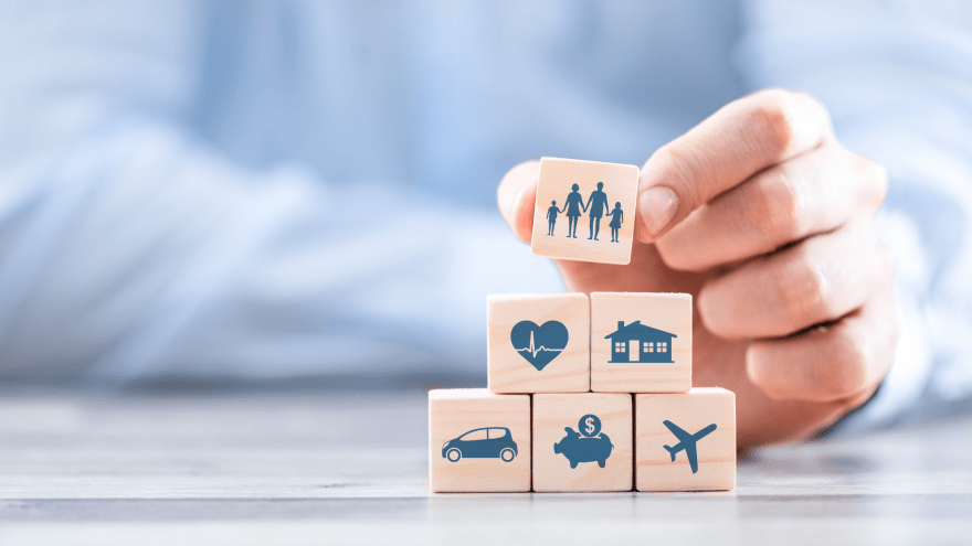 Würfel mit verschiedenen Symbolen (Auto, Sparschwein, Herz, Haus, Flugzeug, Familie) werden zu einer Pyramide gestapelt