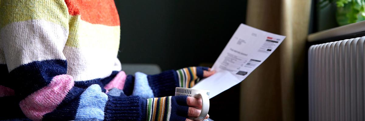 Frau im bunten dicken Wollpulli sitzt mit Teetasse und Heizkostenabrechnung vor einer Heizung