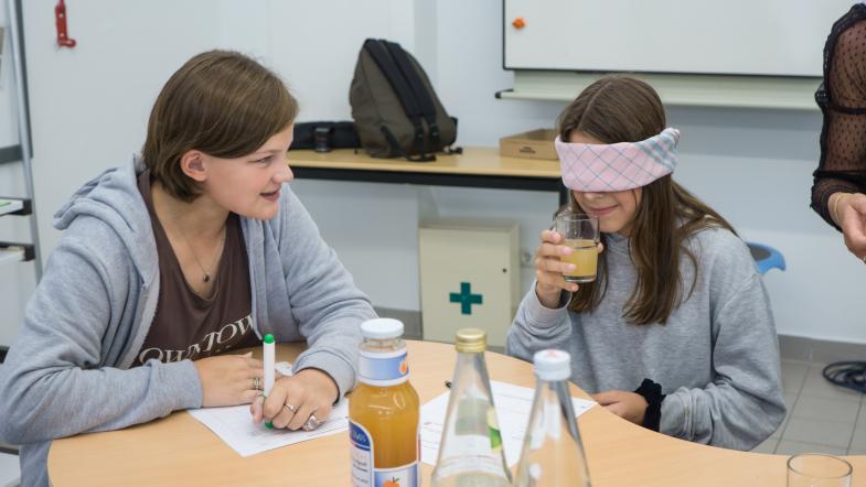Schülerinnen im Klassenraum - Mädchen mit verbundenen Augen probiert Apfelsaft aus einem Becher