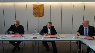 Claus Christian Claussen, Justizminister in Schleswig-Holstein sowie Jochem Schlotmann, Verwaltungsratsvorsitzender der VZSH und VZSH-Vorstand Stefan Bock sitzen an einem Konferenztisch und unterzeichnen eine Vereinbarung.