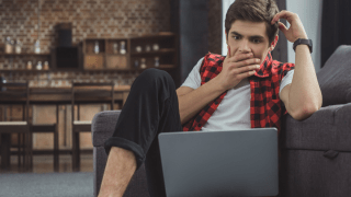 junger Mann auf dem Sofa mit Laptop blickt erschrocken