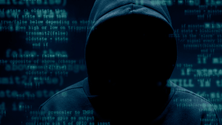 eine Person in dunklem Kapuzenpullover, dessen Gesicht im Dunkeln ist, steht vor eine Wand, auf der Codes laufen