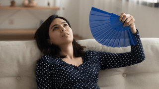 Eine Frau sitzt vor ihrem Sofa und fächert sich mit einem Fächer Luft ins Gesicht.