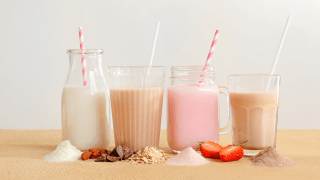 Vier Gläser mit verschiedenen Trinkmahlzeiten stehen auf einem Tisch. Vor den Gläsern liegen die jeweiligen Inhaltsstoffe in Pulverform.