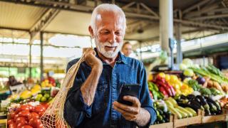 Älterer Herr kauft Gemüse auf Markt ein und guckt lächelnd auf sein Smartphone