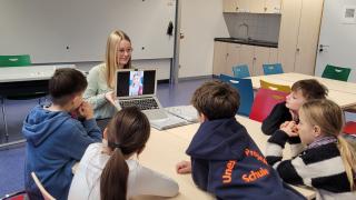 Fünf Schülerinnen und Schüler sitzen an einem Tisch und gucken auf einen Laptop, den eine Studentin der Europa-Universität Flensburg hochhält, eine Spielanleitung an.