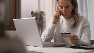 eine Frau sitzt telefonierend vor ihrem Laptop und guckt verunsichert auf ihre Kreditkarte