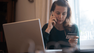 Eine Frau sitzt vor ihrem Laptop,  telefoniert und schaut auf Ihre Kreditkarte