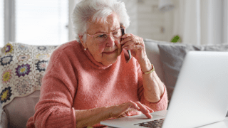 Eine ältere Frau sitzt vor dem Laptop und telefoniert