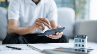 Mann sitzt vor Unterlagen, auf denen ein Modell eines Hauses steht. Er bedient einen Taschenrechner.